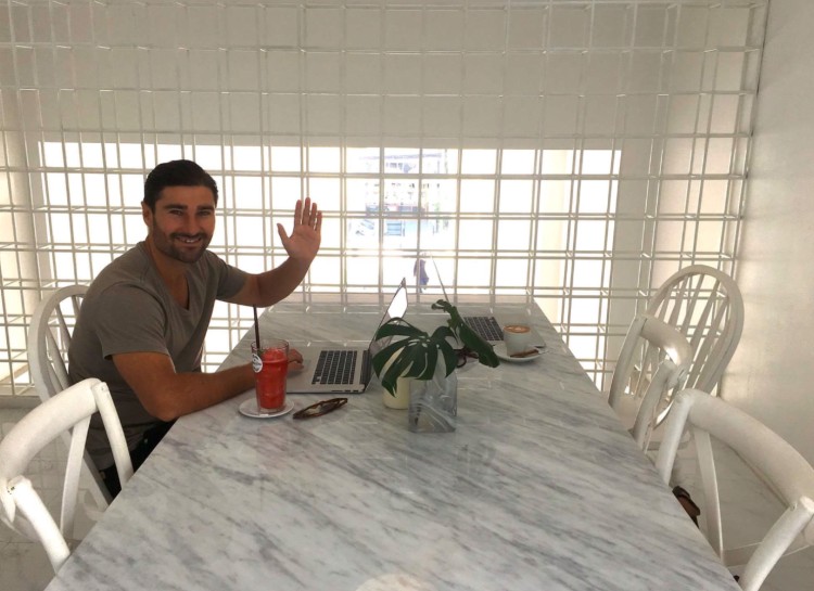 Liam Austin sitting at a marble table in a bright cafe, waving at the camera while working on his laptop to help clients grow their business.