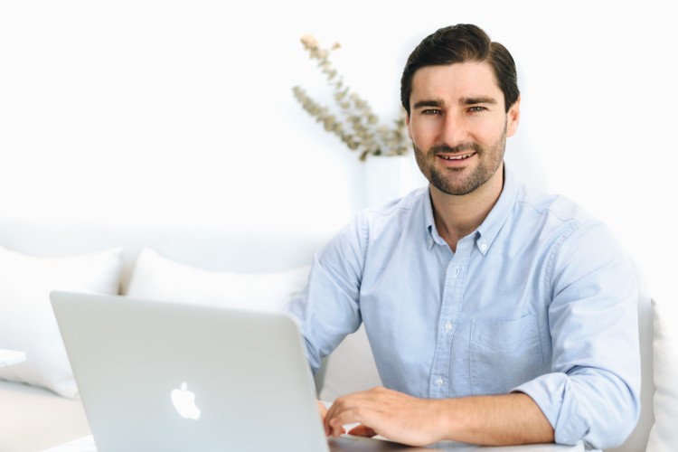 Man in a light blue shirt smiling while working on a laptop, representing a digital coach developing his business strategy.