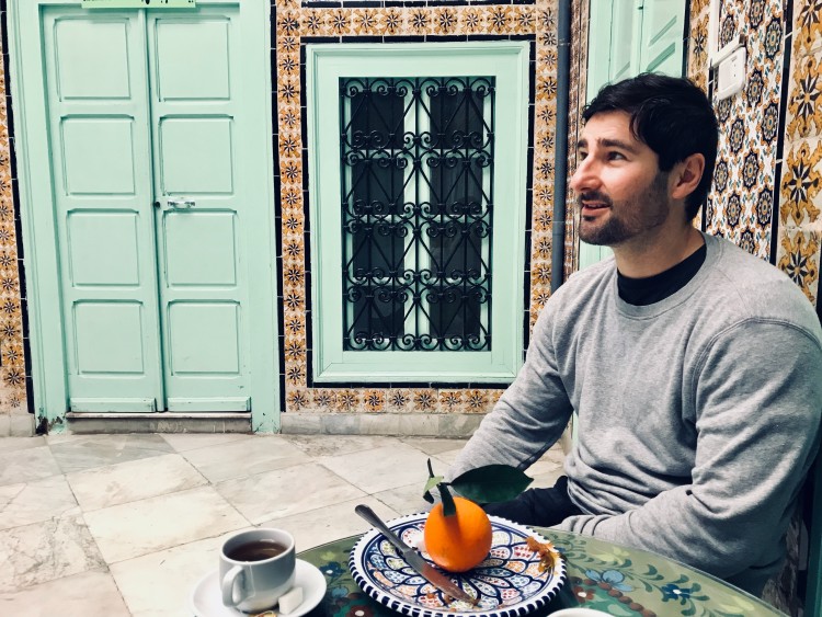 Man sitting at a cafe table with a cup of coffee and an orange, symbolizing a balanced and healthy lifestyle in a relaxed environment.